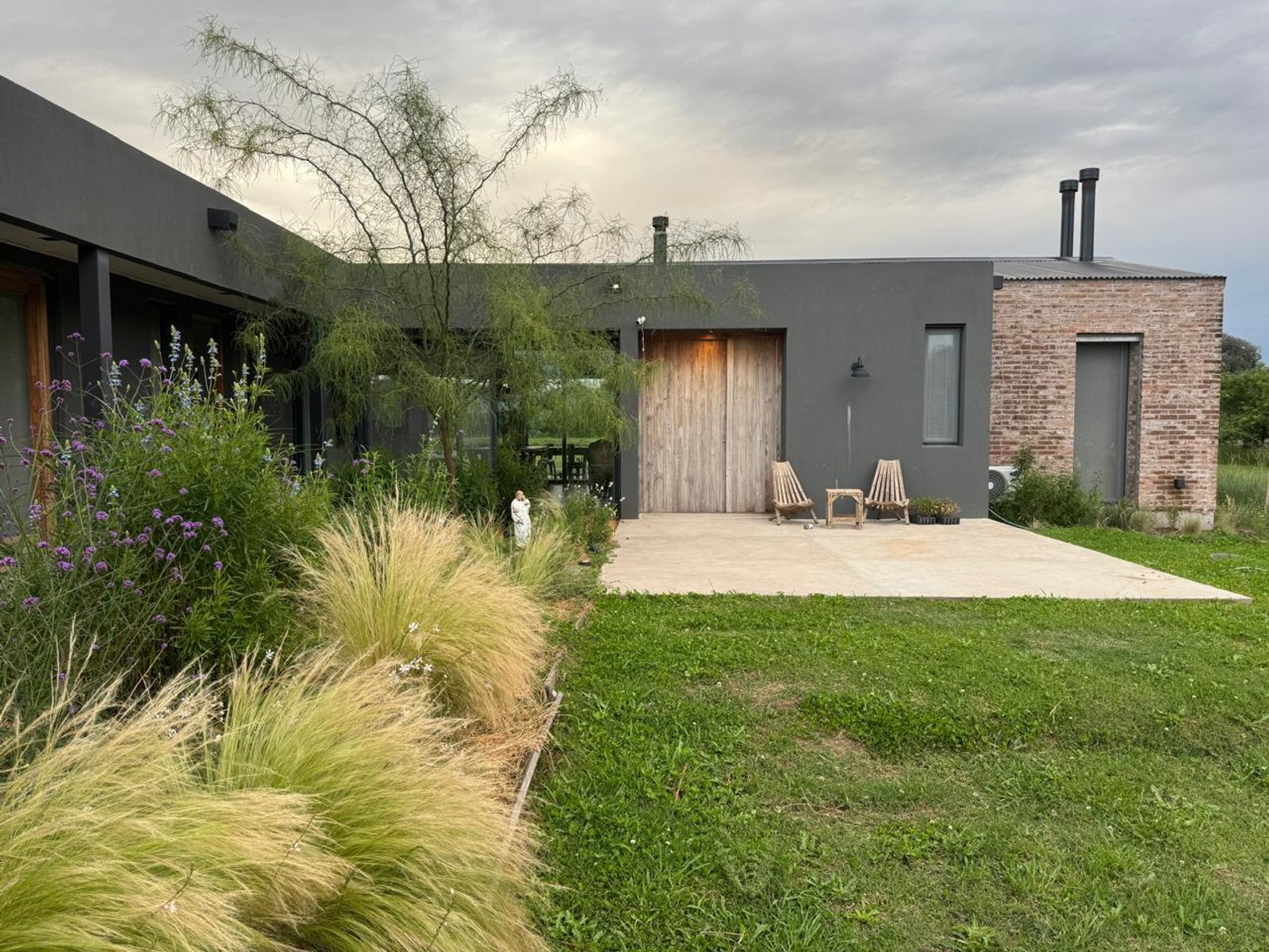 fachada moderna con césped alto y árboles en entorno rural — proyecto Casa Areco, por R+B arquitectos, en San Antonio de Areco